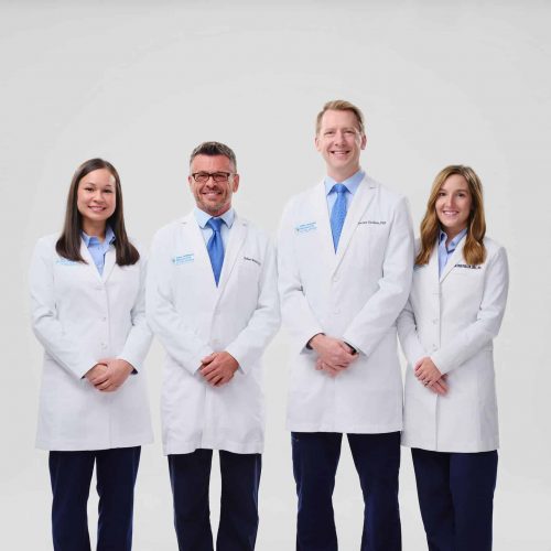 Four oral surgeons from Oral Surgery Specialists of Oklahoma posing together in matching navy branded T-shirts and scrub pants against a white studio background