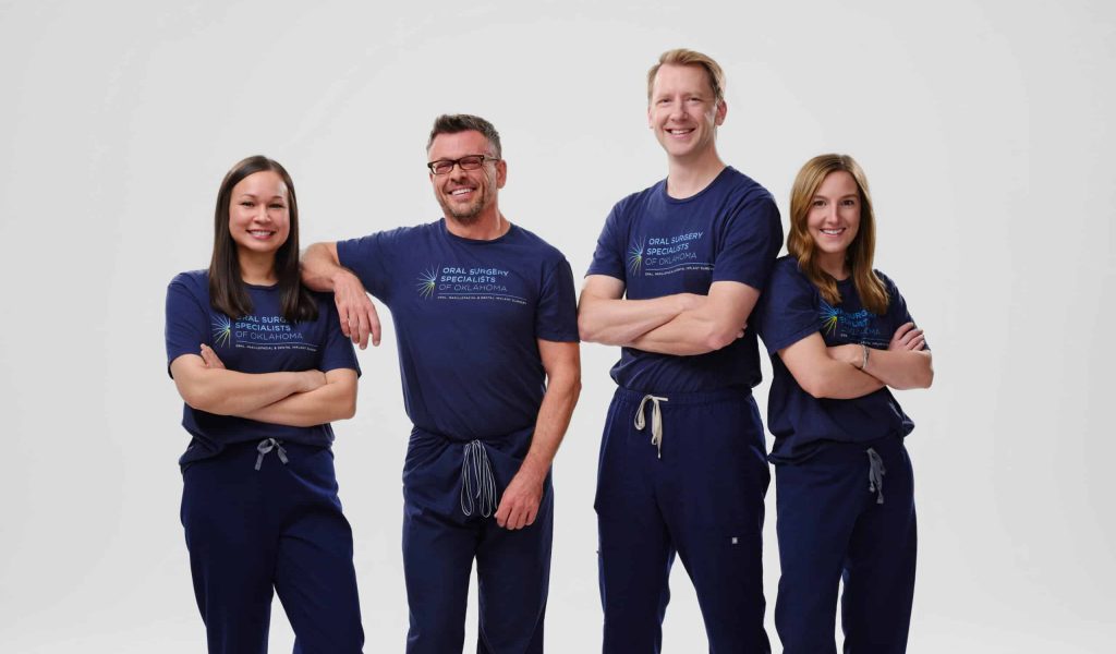 Four oral surgeons from Oral Surgery Specialists of Oklahoma posing together in matching navy branded T-shirts and scrub pants against a white studio background